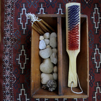 Wooden box with stones, a brush, and a pine cone on a patterned rug.