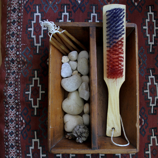 Wooden box with stones, a brush, and a pine cone on a patterned rug.